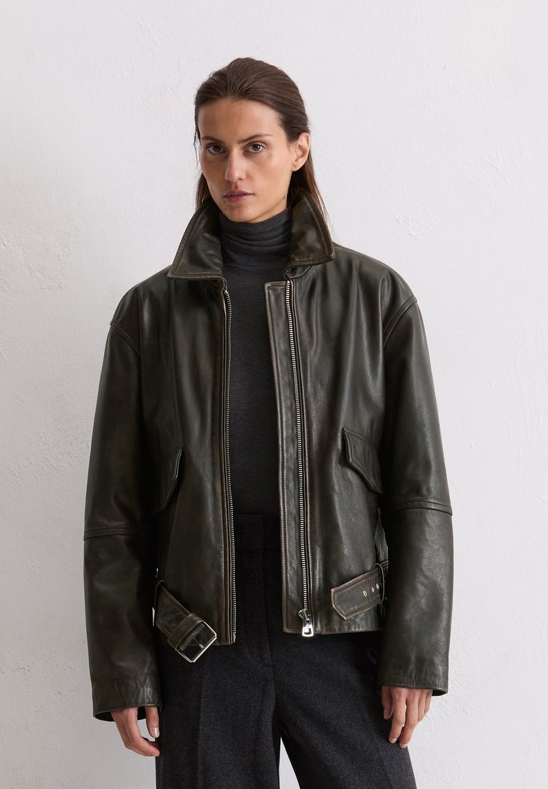 Veste en cuir vert foncé avec un col haut, fermeture éclair à l'avant et poches latérales ; présente un détail de courroie à boucle à la taille.