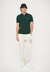 Πράσινο πόλο μπλουζάκι, κρεμ παντελόνι φόρμας με λεπτομέρεια λογότυπου, μαύρο καπέλο και λευκά αθλητικά παπούτσια. Το υλικό φαίνεται να είναι μίγμα βαμβακιού, σε χαλαρή γραμμή, με καθαρό σχέδιο.