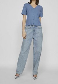 Žena v modré krátké rukávové pruhované košili na knoflíky, v lehkých volných džínách a stříbrných lodičkách s špičatou špičkou, stojící před jednobarevným pozadím.