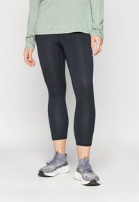 Leggings capri neri con una texture liscia, abbinati a scarpe da ginnastica grigie con un design testurizzato e un sistema di allacciatura. Maglia a maniche lunghe verde.