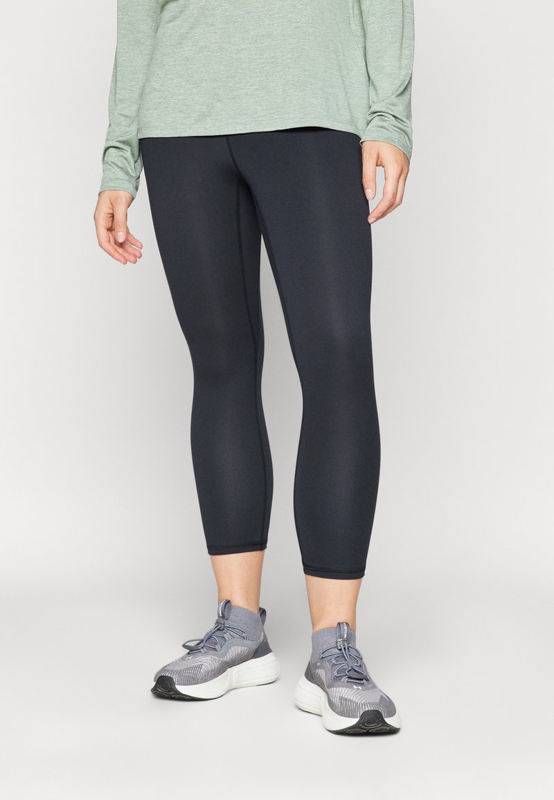 Leggings capri neri con una texture liscia, abbinati a scarpe da ginnastica grigie con un design testurizzato e un sistema di allacciatura. Maglia a maniche lunghe verde.