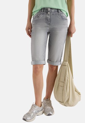 Osoba w podwiniętych szarych szortach jeansowych, zielonym topie, srebrnych sportowych butach, trzymająca beżową torbę na ramię przy boku.