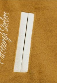 Brauner Filzstoff mit einem weißen Lederdetail mit zwei parallelen Streifen sowie besticktem Text an der Seite.