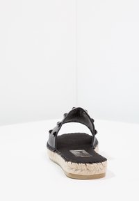 Svart lädersandal med en bred rem, prydd med små nitar. Sula inlindad i jute, texturerad fothydda och slät innersula.