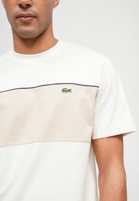 Μπλούζα κοντομάνικη σε κρεμ απόχρωση με μπεζ οριζόντια ρίγα, διαθέτοντας κεντητό πράσινο λογότυπο κροκόδειλου και μαύρες λεπτομέρειες.