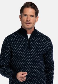 Marineblauwe gebreide trui met een ritskraag, voorzien van een geometrisch lichtblauw patroon. Gemaakt van zacht materiaal met een gestructureerde afwerking.