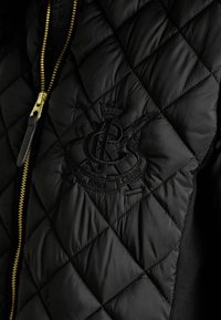 Čierna prešívaná bunda so zlatým zipsom a vyšívaným logom s detailom koruny. Hladká textúra s prešívanými časťami a kontrastnými pletenými stranami.