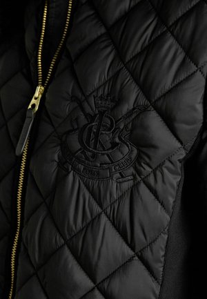Musta tikattu takki, jossa on kultainen vetoketju ja kirjailtu logo kruunudetaljilla. Sileä pinta, jossa on toppatut osiot ja kontrastiväriset neulotut sivut.