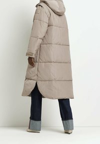 Abrigo acolchado beige con capucha, con un diseño acolchado y aberturas laterales. Combinado con jeans oscuros y acentos de mezclilla remangados.