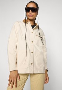 Vrouw met gevlochten haar die grote donkere zonnebrillen draagt, een crèmekleurige knoopjas met een bruin kraag en beige broek, poserende met haar hand op de heup.