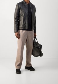 Schwarze Lederjacke mit Stehkragen, schwarzes T-Shirt, beigefarbene Hose und schwarze Sneakers. Hält eine schwarze strukturierte Tasche mit Reißverschluss.