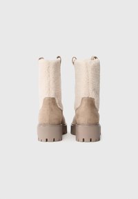 Beige suède laarzen met dikke rubberen zolen en een crèmekleurige shearling schacht, van achteraf weergegeven op een effen witte achtergrond.