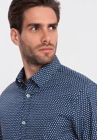 Camicia blu navy con motivo a cuori bianchi, colletto a bottoni e maniche corte. Il tessuto appare liscio con un design aderente e una leggera lucentezza.