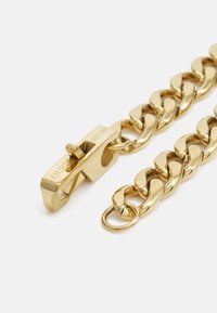 Gullmetallkjede med store lenker, utstyrt med en lås gravert med "GUESS EST. 1981" og en rund ringfeste. Glatt tekstur.
