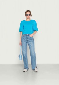 Top azul cortado com mangas curtas bufantes, combinado com jeans de cintura alta em um azul claro com fendas laterais. Bolsa azul clara e óculos de sol.