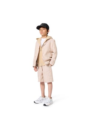 Jongen draagt een beige blazer, bijpassende korte broek, beige hoodie met ritssluiting, witte sneakers en een zwarte baseballpet, staand met handen in zakken.