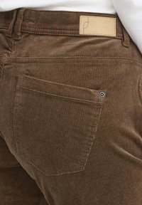 Rudos spalvos korduro kelnės su horizontaliu briaunotu raštu, galinė kišenė su metaliniu akcentu ir virš juosmens esančia firminė odine žyma.