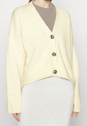 Ivoorkleurig textuur cardigan met een V-hals, verlaagde schouders, vijf grote knopen en een cropped ontwerp, gedragen over een geribbelde beige top.