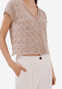 Ljusbeige kort cardigan i öppen stickad mönster, med V-ringning och fem knappar, kombinerad med högmidjade, krämfärgade byxor.