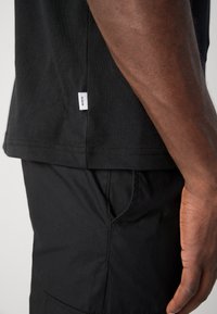 Black cotton t-shirt with a rectangular hem, featuring a small white label on the side. Paired with black pants. Smooth texture.
