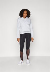Grijze hoodie met witte koordjes, gecombineerd met zwarte legging met gaas-inzetten en zijstrepes, en witte sportschoenen.