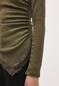 Alyvuogių žalia palaidinė su tekstūruota medžiaga, puošta surinkimu ir nėrinių apačia. Ilgos rankovės, prigludęs dizainas ir subtilūs akcentai.