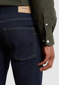 Jeans in denim blu scuro con un'etichetta in pelle marrone che reca la scritta “FARAH”. I jeans hanno un taglio classico con cuciture visibili e una tasca posteriore.