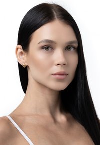 Zelta gredzena auskari ar savīti dizainu, papildināti ar gludu, taisnu melnu matu un neitrāla tonēta ādas krāsu.