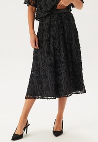 Schwarzer Midirock aus floraler Spitze mit strukturiertem Muster, gewelltem Saum und elastischem Bund, kombiniert mit schwarzen High Heels.