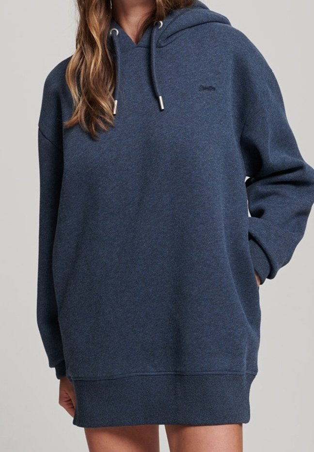 Femme portant un sweat à capuche bleu marine oversize avec cordons, debout avec une main dans la poche sur un fond neutre.