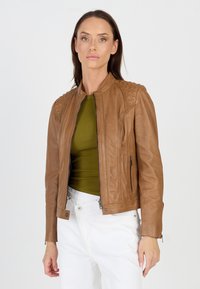 Bruine leren jas met ritssluiting, gestructureerde schouderaccenten en zijzakken. Draagt over een groene top en witte broek.