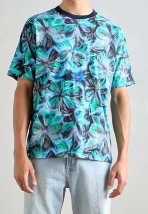 Mężczyzna w krótkim rękawie T-shirtu z abstrakcyjnym wzorem motyla w kolorach niebieskim, zielonym i czarnym oraz w jasnoniebieskich dżinsach.