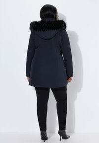 Parka azul marino con capucha de piel sintética, mangas largas y un diseño ajustado. Llevada sobre mallas negras y botines de tacón aguja.