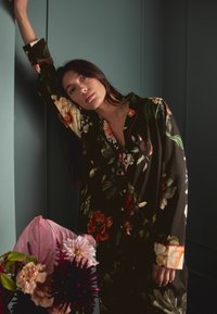 Květinová košile s knoflíky v tmavé tkanině, se silnými, barevnými květy a kontrastními červenými akcenty na límci a manžetách. Růžové květy v popředí.