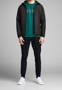 Giacca nera imbottita sopra una felpa verde con testo bianco, abbinata a pantaloni cargo neri e sneaker bianche. Focus su texture e contrasto di colori.