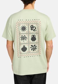 Světlounce zelené bavlněné tričko s velkým grafickým motivem na zadní straně, který zobrazuje opakující se vzor tvarů v černé barvě a s světle růžovými akcenty.