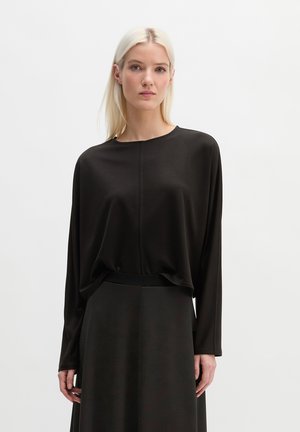 Vrouw met lang blond haar, gekleed in een loszittende zwarte top met lange mouwen en een zwarte rok, staand tegen een effen lichte achtergrond.