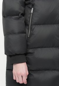 Zwarte pufferjack met een gladde textuur, voorzien van grote geborduurde stukken en een zijzak met ritssluiting. Mouwen in een contrasterende donkere stof.