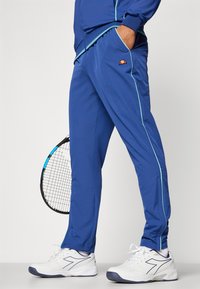 Blaue Sporthose mit türkisfarbenen Seitenstreifen, elastischem Bund und kleinem Logo. Kombiniert mit weißen Turnschuhen.