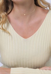 Goldene Choker-Halskette mit einem zarten Herzanhänger, ausgestattet mit einer dünnen Kette und einem minimalistischen Design. Getragen über einem hellbeigen strukturierten Oberteil.
