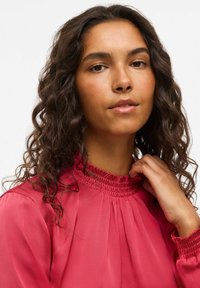 Blusa rosa con scollo arricciato, maniche lunghe e dettaglio plissettato sul davanti. Il tessuto appare morbido e liscio con una leggera lucentezza.