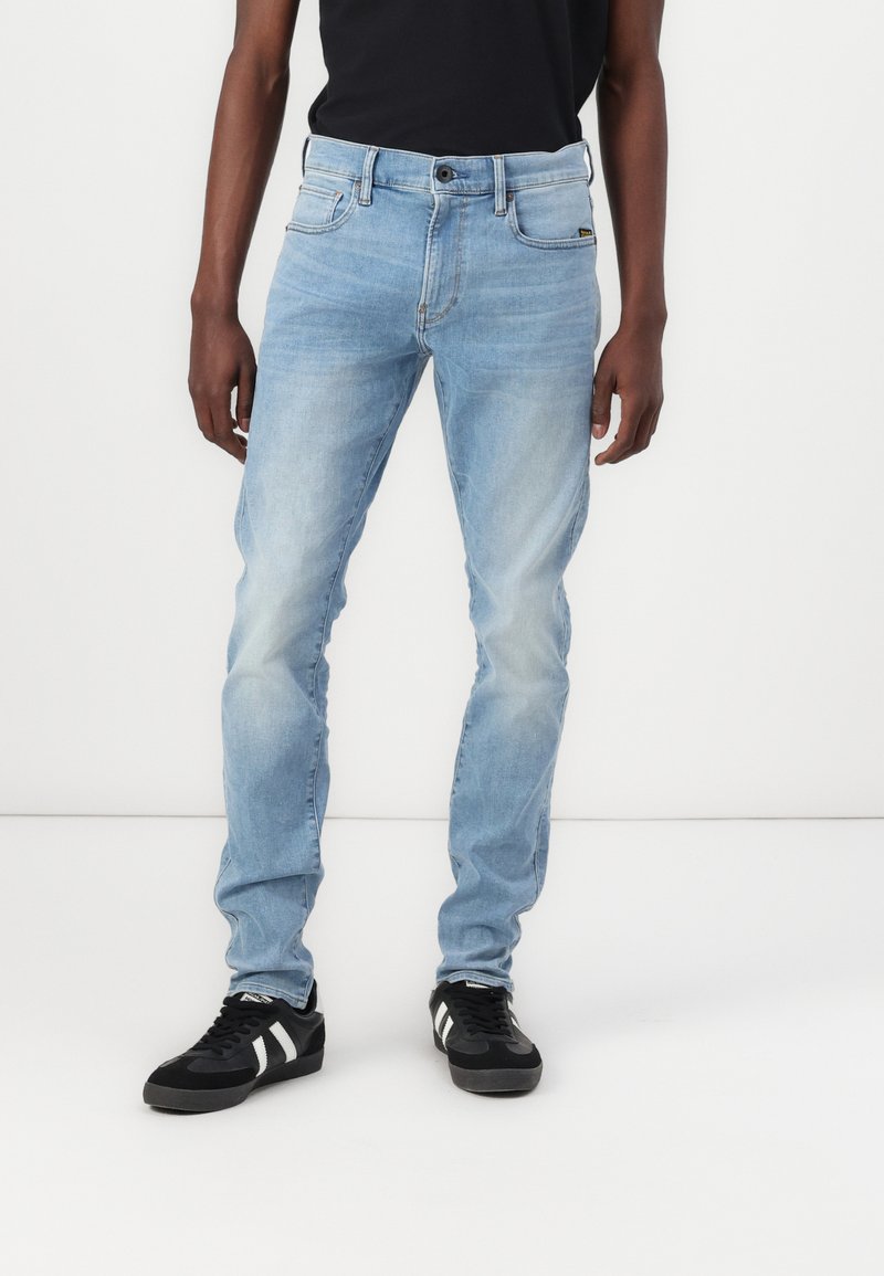 Jeans en denim bleu clair avec une coupe slim, cinq poches et une légère décoloration. Associés à des baskets noires avec des rayures blanches.