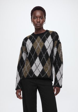 Femme debout portant un pantalon noir à jambes larges, des ballerines noires et un pull à manches longues à losanges noirs, blancs et verts olive.