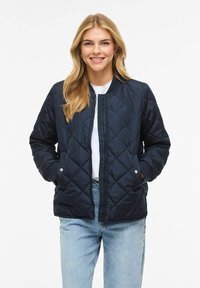 Temno modra prešita bomber jakna z rebrastim ovratnikom, zadrgo in stranskimi žepi. Oblečena prek bele majice s kratkimi rokavi in kombinirana s svetlo modrimi jeans hlačami.