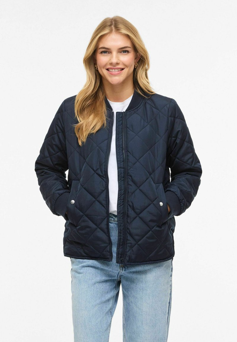 Temno modra prešita bomber jakna z rebrastim ovratnikom, zadrgo in stranskimi žepi. Oblečena prek bele majice s kratkimi rokavi in kombinirana s svetlo modrimi jeans hlačami.