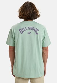 Lichtgroene T-shirt met korte mouwen en een groot paars "BILLABONG" logo en golfafbeelding op de achterkant. Zachte katoenstof, losse pasvorm.