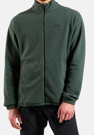 Homme portant une veste polaire zippée vert foncé à manches longues avec poignets élastiques, associée à un pantalon noir.