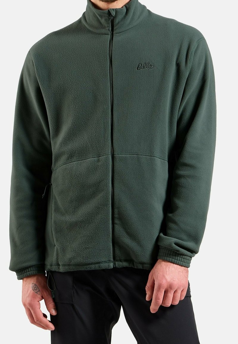 Homme portant une veste polaire zippée vert foncé à manches longues avec poignets élastiques, associée à un pantalon noir.