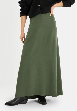 Une personne porte une longue jupe vert olive, des bottines noires et un pull noir boutonné, debout avec une main sur la hanche.