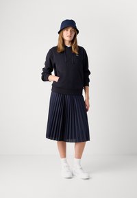 Mujer con un sombrero bucket azul marino, sudadera con capucha azul marino y bolsillo, falda midi plisada azul marino, calcetines blancos y zapatillas blancas, de pie frente a un fondo liso.